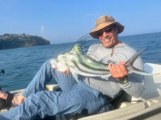 Smiling fisherman sitting on a boat holding up a caught roosterfish with distinctive long dorsal spines