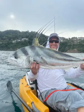 Smiling angler holding a large roosterfish while sitting in a yellow kayak on the ocean