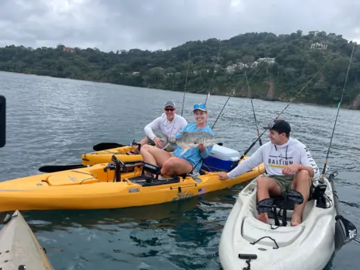 A woman holding up a caught fish while sitting in a yellow kayak on the water alongside two other kayakers