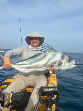 Man in a sun hat sitting in a yellow kayak and holding a large roosterfish on the ocean