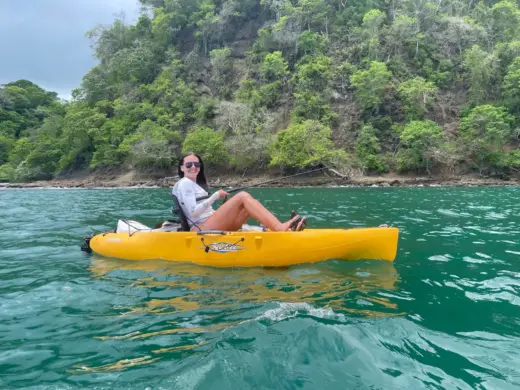 A smiling woman fishing from a yellow pedal kayak in turquoise water near a lush, rocky cliff