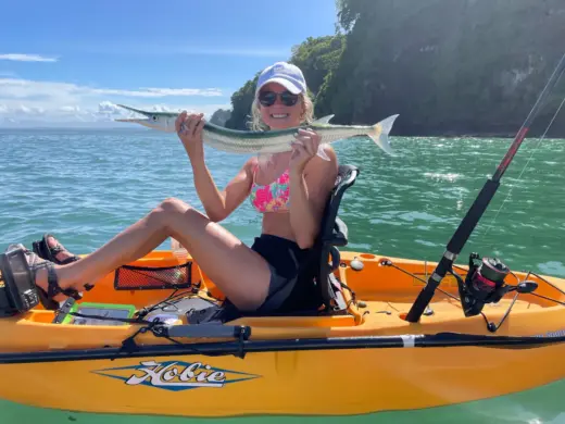 A woman sitting in a yellow kayak on the water holds up a long, slender needlefish she caught