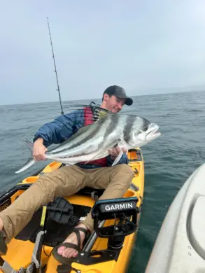 A fisherman sitting on a yellow kayak in open water holds a large roosterfish