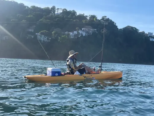 A person wearing a sun hat fishing from a yellow kayak in calm ocean waters near a forested coastline